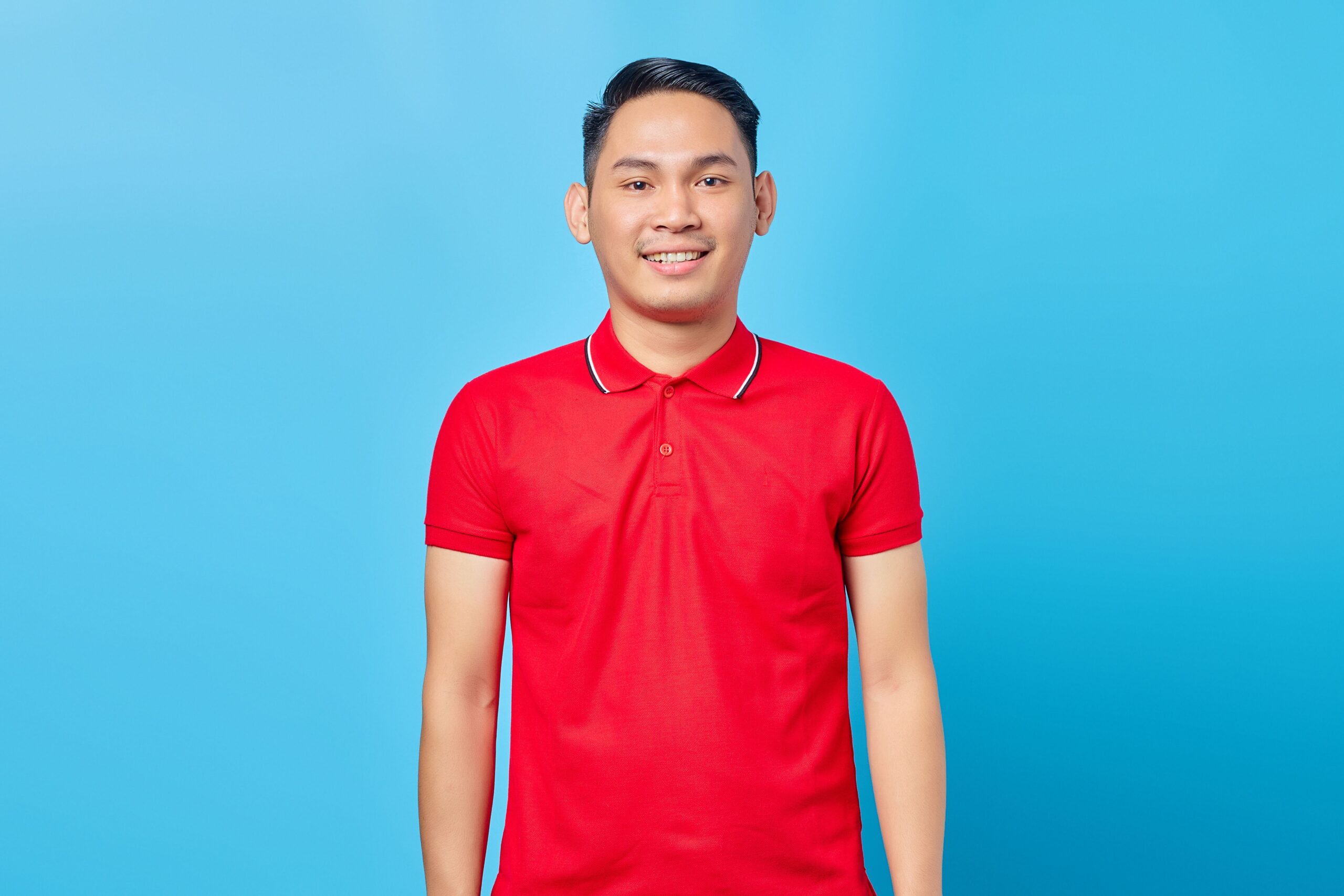 Cheerful young adult man wearing a red polo shirt, posing against a bright blue backdrop.