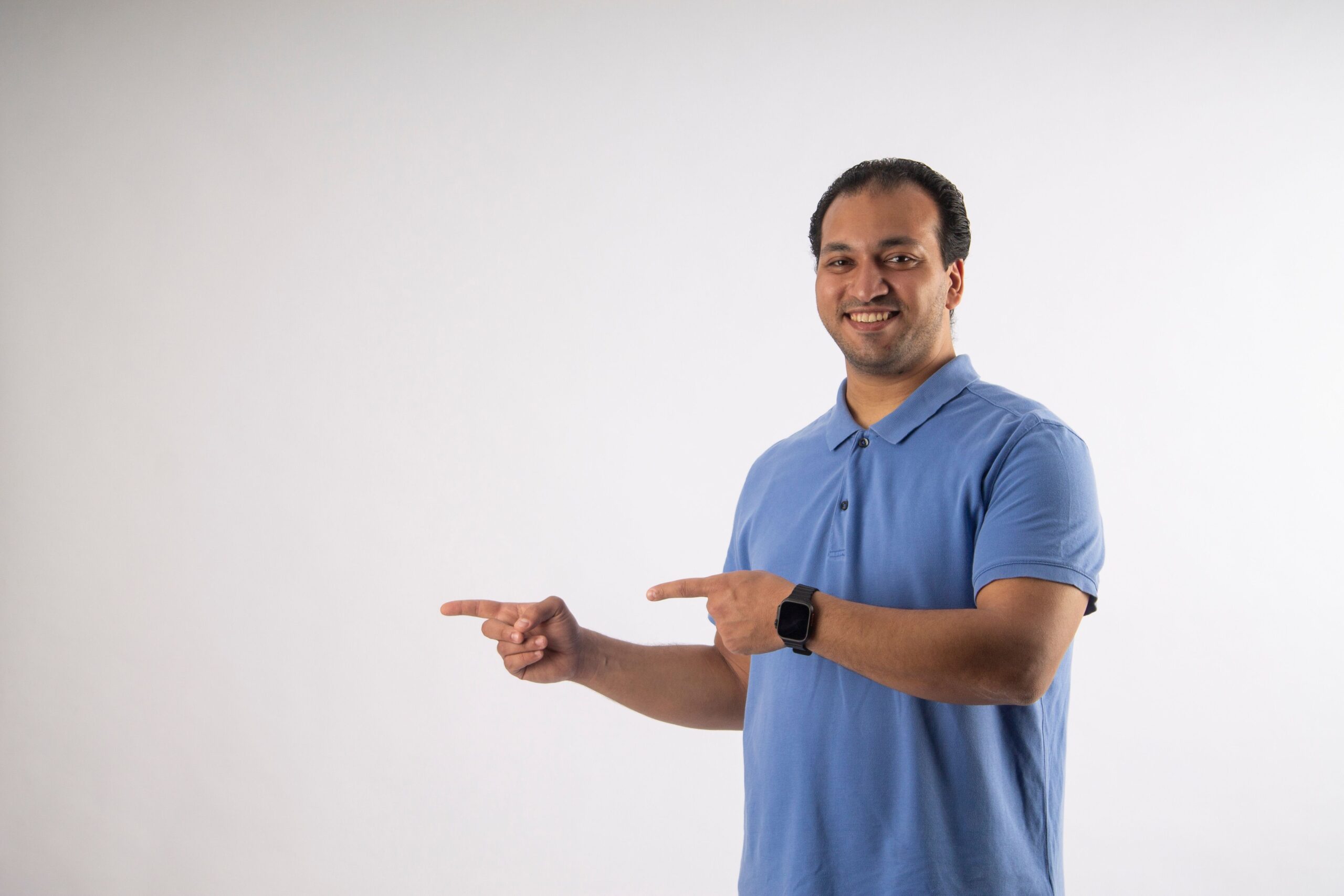 Smiling man in blue polo shirt pointing sideways on blank background.